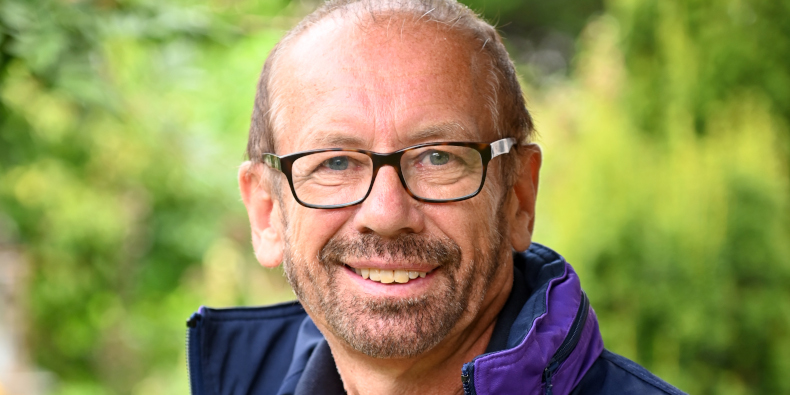 Ein lächelnder Mann mit Brille und Bart, trägt eine dunkelblaue Jacke, steht vor einem verschwommenen, grünen Hintergrund.